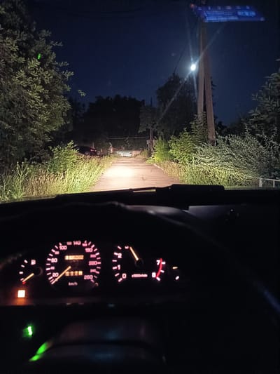 The image is a first-person perspective from inside a car driving on a dark, rural road at night. The road is asphalt with grass and trees lining both sides. In the distance, streetlights illuminate the path. The car's dashboard is visible, showing a speedometer and tachometer, illuminated with pink light. The car's headlights are on, casting a bright beam forward. A street sign is visible on a utility pole on the right side of the road, displaying text and a graphic. The sky is dark blue, indicating it is nighttime. The environment suggests a suburban or rural area. The location is identified as Suklieia, Moldova.