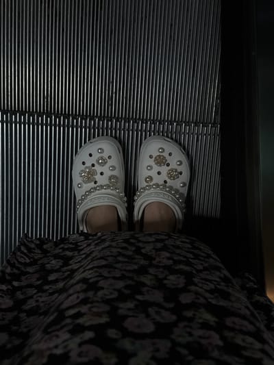 An overhead view depicts the lower body of an individual standing on the ridged metallic tread of an escalator or moving walkway in Kloten, Switzerland. The person is wearing a dark-colored garment, likely a dress or skirt, featuring a subtle light floral pattern. On their feet are white Crocs, extensively adorned with faux pearls of varying sizes and sparkling charms, including square-shaped designs with clear stones and flower-shaped designs with central pearls and clear stones. Each Croc also has a strand of medium-sized faux pearls draped over its upper strap. The escalator surface comprises numerous parallel, silvery-gray metallic ridges, flanked by dark, unreflective side panels. The overall lighting is dim, casting a subdued glow on the reflective elements of the footwear and the escalator treads.