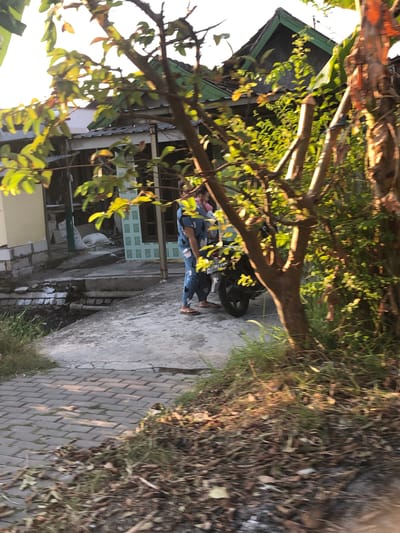In Putat, Indonesia, a person in blue pajamas with a white and black pattern is standing next to a motorcycle. Another person, possibly a child, is partially visible behind them, being held by the first person. The motorcycle is dark colored. They are on a paved area next to a small canal with visible pipes. Behind them, there is a building with a green roof and a wall constructed of white blocks. There are trees and foliage in the foreground and background, with sunlight filtering through the leaves. The ground in the foreground is paved with bricks and covered with dry leaves and grass.