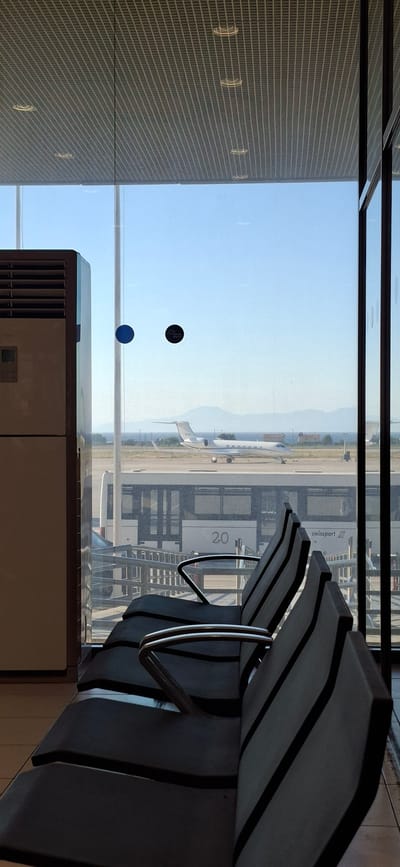 The image depicts an airport lounge with a view of an airport tarmac. In the foreground, rows of dark, modern airport seating are visible, angled towards a large window. The window offers a clear view of the outside, revealing an airplane on the tarmac and a distant landscape of hills under a bright, clear sky. The lighting suggests it is daytime, possibly late morning or early afternoon, with the sun casting distinct shadows.
On the window, there are two circular stickers; one is black with a starburst logo, and the other is a solid blue circle. Beyond the plane, there's a building with the text "SWISSport" and "2.0" visible on its side, indicating an airport service provider. The presence of airport seating, an airplane, and a building labeled "SWISSport" strongly suggests the setting is an airport terminal. The weather appears to be sunny and clear, with no visible clouds. The scene conveys a sense of waiting and transit, common to airport environments. The overall impression is one of functional modern design and clear visibility, typical of contemporary airport lounges. The location is identified as Rhodes, Greece, which aligns with the possibility of clear, sunny weather.