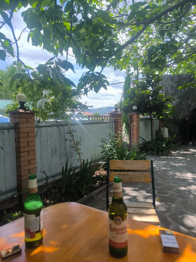 The image depicts an outdoor scene in Karatakla, Georgia. A wooden table is prominent in the foreground, holding two partially consumed beer bottles, one green and one Budweiser, and a pack of cigarettes. A simple wooden chair sits behind the table. Beyond the table, the scene opens to a patio area paved with irregular stone slabs. A low stone wall with brick pillars marks the edge of the property, beyond which glimpses of trees, buildings, and a distant mountain range are visible under a partly cloudy sky. Lush greenery surrounds the patio, with plants and trees providing shade. A figure in dark clothing is seen in the background, near the stone wall.