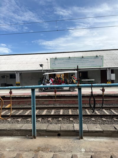 The image depicts a railway station in Thanjavur, India, under a cloudy blue sky. In the foreground, a platform runs alongside train tracks. A blue metal railing, partially rusted, separates the platform from the tracks. On the platform, a small electric vehicle, resembling a golf cart, carries several passengers. The passengers are dressed in casual attire, with one individual wearing a red and white outfit. Behind the vehicle, a building with a white, corrugated metal roof stretches across the frame. A sign above the building reads "INDIAN RAILWAY CATERING AND TOURISM CORPORATION LIMITED (A GOVERNMENT OF INDIA ENTERPRISE - MINI RATNA) REFRESHMENT ROOM (KITCHEN UNIT) THANJAVUR JUNCTION". The environment appears to be a functional, outdoor setting at a transportation hub.