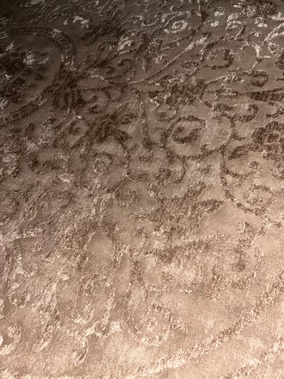 This is a close-up, overhead shot of a carpet or rug with an ornate, swirling pattern. The pattern is in a darker brown against a lighter tan background. The lighting casts shadows and highlights on the textured surface, giving it a slightly fuzzy appearance. There are no people, objects, or discernable environmental features that would indicate a specific location in Ponebel, Ukraine, or any other city.