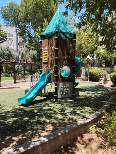 The image shows a children's playground located in Zikhron Yaakov, Israel. In the foreground, there is a blue slide with a rope net below it, attached to a multi-level play structure. The play structure is primarily brown with turquoise accents on its peaked roof. The ground cover appears to be artificial turf, with dappled sunlight filtering through the surrounding trees. In the background, there is a wooden pergola with a bench underneath, and beyond that, buildings and more greenery. A lamppost stands to the right of the play structure. The overall scene depicts an outdoor recreational area on a sunny day.