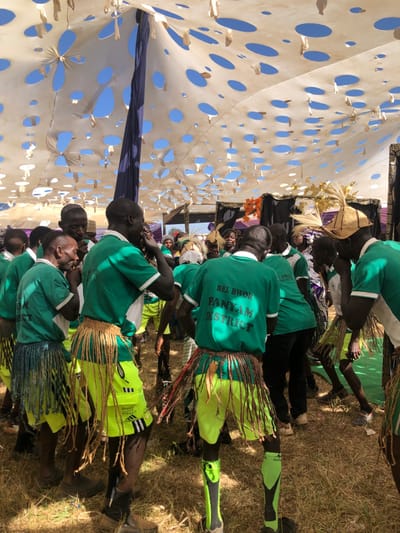 A vibrant outdoor cultural event is captured from a slightly elevated perspective, with the image rotated 90 degrees counter-clockwise. The scene is set under a large, light-colored canopy perforated with numerous circular cutouts, revealing a bright blue sky above. Small, light-colored fabric pieces protrude from some of the cutouts, adding texture. A dark blue or purple fabric section is visible deeper within the covered area.
The foreground and midground are filled with a group of primarily males, many of whom appear to be young adults, dressed in matching green short-sleeved shirts, some with white trim. Several individuals wear yellow or lime green shorts or trousers, often adorned with traditional-looking waist wraps or skirts made of braided grass or raffia material in brown, yellow, and green tones. Some participants wear elaborate headwear, including woven straw hats decorated with feathers or straw accents. One person wears lime green football socks and possibly athletic shoes. The ground beneath them is dry earth with sparse patches of grass, indicating an outdoor setting in Kerang, Nigeria.
The participants are mostly seen from behind, suggesting they are part of a performance or procession. The backs of their green shirts bear white text: "BEL BWOR" on the top line, "PANYAM" on the middle line, and "DISTRICT" on the bottom line. The bright sunshine suggests it is daytime, likely midday or early afternoon. The gathering appears to be a festive or ceremonial occasion, possibly a cultural dance or community event.