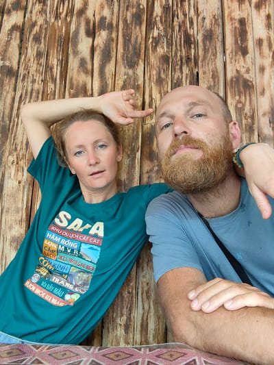 A medium, eye-level shot depicts two adults seated indoors, positioned against a rustic wooden wall.
The woman, fair-skinned with blonde hair, is wearing a teal t-shirt with multiple colorful images. She has one arm raised, hand near her head. Her expression is neutral.
The man, fair-skinned and bald with a reddish beard, is wearing a light blue t-shirt. He is slightly in front of the woman. He has one arm resting on the table in front of him and the woman's hand on his shoulder. His expression is a small frown.
The wooden wall consists of vertical planks.
The bottom of the frame shows a patterned tablecloth, likely covering a table.
The t-shirt has the following text: Sapa KHU DU LỊCH CÁT CẮT HÀM RỒNG LỢN MVR CẮP MAVERICK NÁCH THỊT TRÂU GÁC BẾP COKÍNH THANG XÔI BẢY MÀU VƯỜN QUỐC GIA HOÀNG.
Given the text "Sapa," it is inferred the photograph was taken in the city of Thành phố Hà Giang, Vietnam.