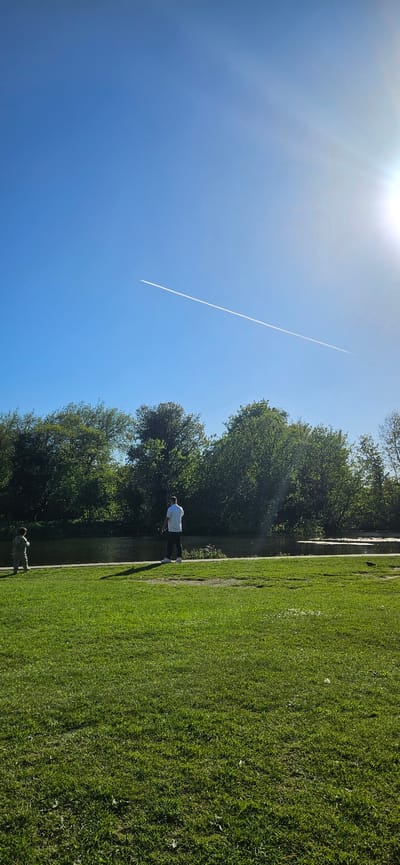 This image, correctly oriented, depicts a serene outdoor scene, likely a park or canal bank, bathed in bright midday sun. The upper portion of the frame is dominated by a clear, deep blue sky. The sun is prominently visible in the upper left, radiating a strong glare and lens flares across the sky. A long, thin white contrail from an aircraft cuts diagonally across the blue expanse, extending from the middle left towards the upper right.
Below the sky, the landscape features a vibrant green grassy area that stretches into the foreground and slopes gently towards a calm body of water, possibly a river or canal. The grass appears well-maintained. On the opposite bank of the water, a dense line of mature trees with rich green foliage forms a verdant backdrop, suggesting a lush, natural environment. The water surface shows reflections of the sky and trees.
Two individuals are present in the scene. An adult figure, dressed in a white shirt and dark trousers, stands by the water's edge in the mid-ground, facing towards the water. Another smaller figure, possibly a child, is visible closer to the foreground, also near the water, though partially obscured. Both appear to be observing the surroundings in a relaxed manner. The strong sunlight and clear skies indicate a bright, sunny day, likely during midday or early afternoon, contributing to a peaceful and pleasant atmosphere. The location context provided indicates this scene is in Norwich, United Kingdom. There is no visible text in the image.