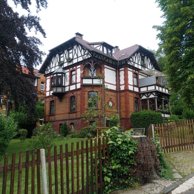 This image shows a two-story brick and half-timbered house in Eisenach, Germany. The house has a red tile roof with dormer windows. The facade features red brick on the lower level and white plaster with dark wood beams on the upper level. A bay window protrudes from the corner of the building on the first floor. A balcony with a white railing and a translucent awning is present on the second floor. The house is surrounded by greenery, including trees, bushes, and a lawn. A wooden fence runs along the front of the property. A large tree stump with ivy growing around it is visible in the foreground, to the right of the fence. The sky is overcast.