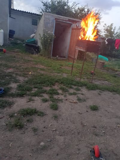 In Pryvilne, Ukraine, an outdoor scene captures a burning barbecue with a tall, vibrant flame engulfing the contents. The barbecue sits in front of a dilapidated, open-door structure with a weathered wooden door. To the left, a light-colored building with a sloping roof is visible, alongside several large blue barrels. The ground is a mix of dirt and sparse vegetation. In the background, trees and a cloudy sky complete the environment. The image appears to be taken during daylight hours, possibly in the late afternoon or early evening, given the lighting conditions.