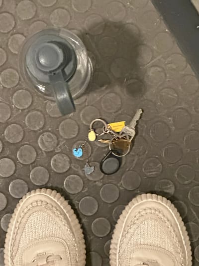 This is an overhead, close-up, forensic photograph of a gym floor with a water bottle and a set of keys. The floor has a pattern of raised circles. On the left of the image is a gray water bottle with a gray lid and a gray strap. In the center of the image are a set of keys on a keyring with several charms. The charms include a yellow oval, a blue bird, and a gray chicken. There are also a few keys, one of which has a yellow tag. The right side of the image shows the top of two light-colored sneakers. The scene appears to be indoors. The image was taken in Lynnwood, United States.