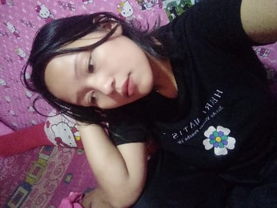A young woman with dark hair wearing a black t-shirt is angled towards the camera, resting her head on her hand. The t-shirt features a white flower design. She has fair skin and appears to be looking down. The background consists of a pink wall and bedspread covered in Hello Kitty patterns. The lighting is soft, giving the image a relaxed and slightly blurred effect.