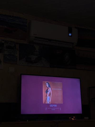 This image depicts a dimly lit indoor environment, identified as a room within Varbak, Bulgaria. Positioned high on a textured wall is a white FINLUX split-system air conditioner, displaying the temperature "22" in blue digital numerals. Directly below the air conditioner, a black-framed flat-screen television is active, showcasing a music player interface. The screen prominently features album art for Toni Braxton's album "The Heat," with the track "He Wasn't Man Enough" by Toni Braxton also displayed alongside music playback controls. The wall space surrounding and above the television and air conditioner is adorned with several dark posters or prints, predominantly featuring various automobiles, including a partial "VW Club" emblem visible on the right side. On the surface immediately in front of and below the television, a gaming mouse with orange illuminated accents is situated on the right, next to a small, circular white object. No individuals are present within the scene, which is primarily illuminated by the light emanating from the television screen and the air conditioner's digital display.