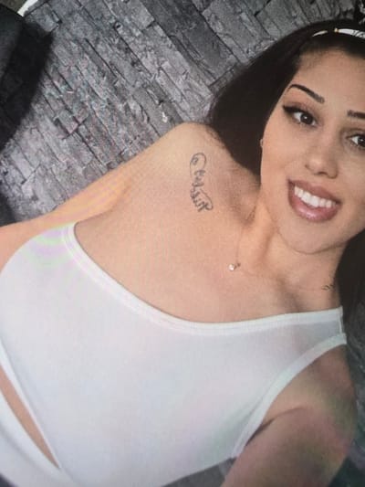 A young woman is prominently featured in a close-up, slightly angled portrait shot, set against a textured, gray brick-patterned wall. She has long, dark hair, pulled back by a white headband, and is smiling broadly, revealing her white teeth. Her eyes are open and looking towards the camera, with visible eye makeup. She wears a white, ribbed, one-shoulder top. A delicate chain necklace with a small, round, clear pendant adorns her neck. On her left shoulder/collarbone area, there is a small tattoo written in script lettering. While partially obscured and stylized, it appears to be a name or word, possibly "Melissa" or a similar term, beginning with an ornate initial. The woman's expression conveys happiness and confidence. The setting is indoors, with the background providing a subtle textural element behind the sharply focused subject. The overall impression is one of a cheerful and candid moment, taking place in Bremerhaven, Germany.