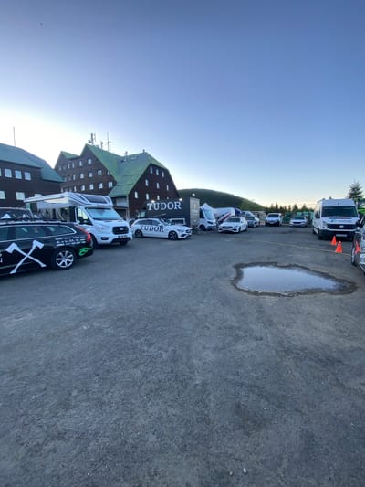 The image shows a parking lot with various vehicles in front of a building with a dark brown facade and a multi-tiered green roof. The word "TUDOR" is displayed on the building.
Several vehicles are visible in the parking lot, including vans and cars. Some vehicles have branded wraps. There is a large puddle on the asphalt surface of the parking lot. The sky is clear and blue. The building's architectural style suggests a location in the Czechia region, and according to the image metadata, the location is in Bělá pod Pradědem, Czechia.