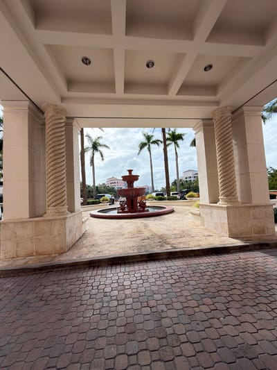 This image shows a view through an arched entranceway in Key Biscayne, Florida, USA. The entranceway is framed by two decorative columns with a spiral design, supporting a coffered ceiling with recessed lighting. Beyond the entrance is a courtyard with a tiered fountain in the center, surrounded by palm trees and other greenery. In the background, buildings are visible. To the right of the entrance is a paved walkway with a patterned brick design. The weather appears partly cloudy, with the sky visible between the palm trees.