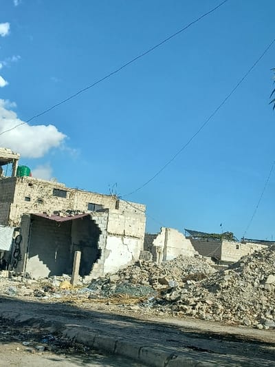 The image displays a scene of extensive urban destruction in Al-Qadam Municipality, Syria, under a clear blue sky with scattered white clouds. In the foreground, a dark asphalt road transitions to a damaged concrete curb littered with debris. Directly beyond the curb, a multi-story building constructed of light-colored concrete blocks exhibits severe structural damage, with a substantial section of its front wall missing, exposing an internal concrete support column and a partial red-brown corrugated roof structure. A green water tank is visible on a rooftop to the left, adjacent to this damaged building. To the right, a large mound of mixed rubble, composed of broken concrete, rebar, and greenish glass fragments, dominates the midground, extending into the background and obscuring further damaged structures. Additional partially collapsed buildings are visible in the distance, one featuring a dark, possibly tarp-covered roof. Overhead power lines traverse the sky. No individuals or active events are depicted.