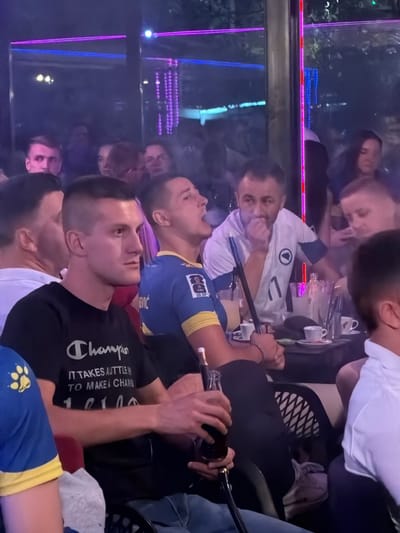 The image depicts an indoor scene, possibly a cafe or lounge, with several individuals seated around tables. The environment is dimly lit, with neon lights in blue and purple colors illuminating the background. In the foreground, three men are prominently visible. The man on the left wears a black t-shirt with a "Champion" logo and text. The man in the center wears a blue and yellow jersey with a FIFA logo, and he is holding a hookah pipe, appearing to exhale smoke. The man on the right wears a white t-shirt with a crest. Several tables are present, with drinks, cups, and an ice bucket on them. The overall atmosphere suggests a social gathering. The image is identified as being taken in Travnik, Bosnia and Herzegovina.