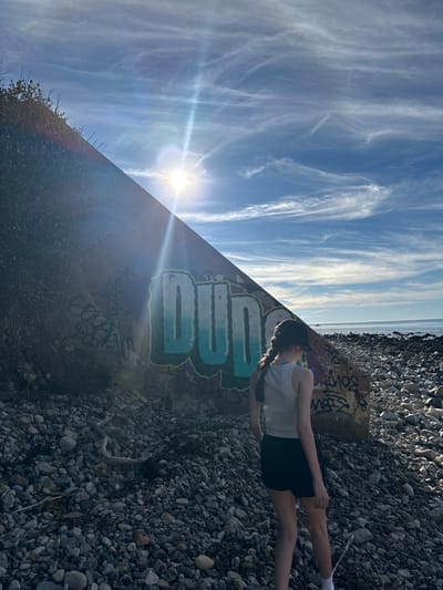 The image depicts a coastal scene in Palos Verdes Estates, United States. A lone female figure, viewed from behind, stands on a rocky beach composed of numerous grey and tan pebbles. She wears a light grey tank top, dark shorts, white socks, and a dark baseball cap over her dark hair, which is styled in two braids. The subject faces right, looking towards the Pacific Ocean, which stretches to the horizon under a clear blue sky with wispy white clouds.

To the left of the figure and occupying the left half of the frame is a large, angled concrete structure, possibly part of a seawall or bunker. Its upper left portion is covered with dark green vegetation. The visible concrete surface is heavily graffitied. The most prominent graffiti is the word "DUDE" rendered in large, blue-green bubble letters with a yellow outline, positioned centrally on the structure. Below and to the right of "DUDE," additional black graffiti text is present, partially illegible. The sun is visible in the upper left quadrant, creating a prominent lens flare that streaks across the upper-left diagonal of the image. The overall environment suggests a rugged, outdoor coastal area with evidence of human activity (graffiti).