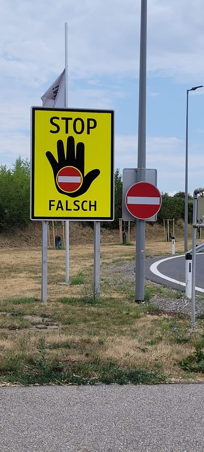 The image depicts a road-side scene in Pöchlarn, Austria, under a partly cloudy sky. A prominent yellow rectangular sign with a black border reads "STOP" in large black letters at the top. Below "STOP" is a black silhouette of a hand with an open palm. Within the palm, a circular sign with a red border and a white horizontal bar in the center, enclosed by a smaller red circle, is depicted. Below the hand is the German word "Falsch" (meaning "False" or "Wrong") in black capital letters. To the right of this sign, a smaller, standard red circular "no entry" sign with a white horizontal bar is mounted on the same pole. The signs are situated on a grassy embankment next to a paved road with a white lane marker. In the background, there are trees and some smaller planted saplings, along with a light pole.
