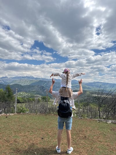 A medium shot shows a person facing the sky, with their back toward the camera. The setting appears to be a mountaintop overlooking a valley in Gornje Mrke, Montenegro. The person is wearing blue jean shorts, a light colored top, and white tennis shoes. They have a backpack on and a pink hat with a blonde braid coming out of it. The person's arms are raised. The sky is filled with white, puffy clouds with patches of blue sky in between. In the background, a mountain range stretches across the horizon. In the foreground, there is a patch of grass with some trees and shrubs. A wire fence is visible in the middle ground.
