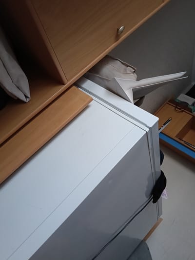 The image shows a close-up view of a white appliance, possibly a refrigerator or a filing cabinet, in an indoor setting. The appliance has a clean, modern design with smooth surfaces and sharp edges. A wooden shelf or cabinet is visible above and to the left of the appliance, with a silver drawer pull. A folded piece of light-colored fabric, possibly clothing or bedding, is tucked between the appliance and the wooden cabinet. A white paper object, possibly a large envelope or a folded piece of cardboard, is partially visible emerging from the top of the appliance. A black fabric item is hanging from the side of the appliance. In the background, a wooden desk or cabinet with blue accents is visible, with some items on its surface, including what appears to be a pen. The flooring is a light-colored linoleum or tile. The scene appears to be located in a residential or office space. No specific details indicate the location in Satbayev, Kazakhstan.