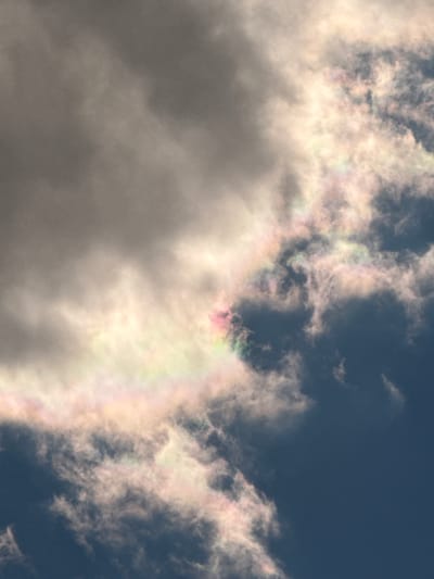 The image shows a cloudy sky. In the upper right of the image, the sky is a dark blue. Most of the image is covered in gray and white clouds. In the center of the image, a patch of iridescent clouds shows bands of color including pink, green, and yellow. There is no information in the image to confirm the location as Hartavan, Armenia. There are no people or objects visible in the image.
