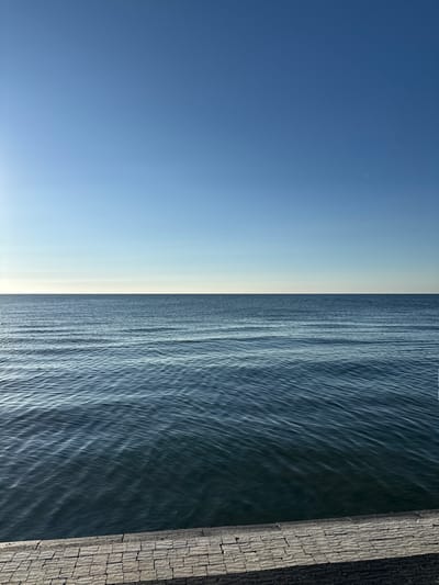 A wide shot captures a tranquil seascape with a clear blue sky meeting the calm sea at the horizon. The foreground features a paved walkway made of rectangular stone blocks. The water exhibits subtle ripples, reflecting the sunlight. The location is the waterfront in Süleymanpaşa, Turkey.