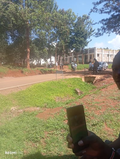 The image captures an outdoor scene, likely a campus or institutional ground, under a clear, bright sky. The setting features lush greenery with numerous tall trees providing shade, reddish-brown soil, and a paved road that curves through the middle ground.
In the foreground, the partially visible profile of a person on the right holds a black smartphone, with its screen reflecting the surroundings. This person also wears a beaded wristband, possibly a traditional Kenyan accessory.
The middle ground is populated by several groups of individuals, possibly students or staff, gathered near the roadside and in front of buildings. They appear to be engaged in casual conversations or waiting. A white car is parked or stopped on the right side of the road near one of these groups.
In the background, modern-style buildings with light-colored facades and structured designs are visible, set amidst more trees. These buildings suggest an educational or administrative complex. A small white signpost stands near the edge of the road in the center.
The scene depicts a sunny day, likely morning or early afternoon, with adequate lighting and a blue sky overhead. The presence of verdant trees, reddish soil, and specific architectural styles aligns with the provided context of Nkomo ward, Kenya. The bottom left corner of the image displays a "REDMI 13C" watermark, indicating the camera used. The overall atmosphere appears calm and routine.
