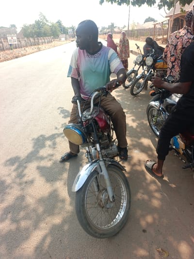 This image captures a sunlit street scene in Nassarawan Eggon, Nigeria. The location is confirmed by a prominent white sign with red lettering for "GENERAL HOSPITAL NASSARAWA-EGGON" visible on the left side of the background.
In the foreground, an adult male with dark skin, wearing a multi-striped t-shirt (white, light blue, pink, purple) and brown trousers with dark sandals, is seated astride a red and silver motorcycle. He is gripping the handlebars and looking towards his left. The motorcycle features a large, round, yellow-tinted headlight with a blue rim and a silver front fender. To his immediate right, a second adult male, partially visible, wears a patterned long-sleeved shirt and dark joggers with dark slides, standing beside a black motorcycle. Further into the midground on the right, a third adult male, wearing dark clothing, is partially obscured while leaning over another motorcycle. Multiple motorcycles are parked or positioned along the dirt shoulder of the paved road.
In the background on the left, two women, dressed in traditional head coverings and long patterned garments (one deep red, one multicolored floral), are standing near a low concrete wall. The paved road surface shows distinct shadows cast from unseen overhead foliage. The overall environment appears dry and semi-urban, under clear daylight conditions.