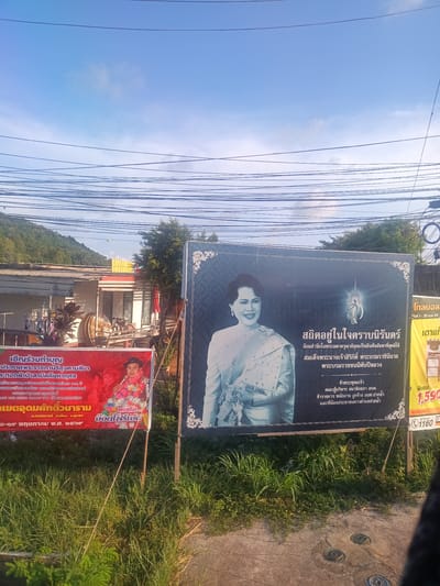 The image shows a roadside scene in Pak Nam Chumphon, Thailand, with two large banners. The banner on the right is a memorial to Queen Sirikit, depicting her in traditional Thai dress, with text in Thai expressing reverence and dedication. The banner on the left is a promotional poster for a charity event at the Khet Udomsak Diwaram Temple. It invites people to participate in a donation campaign. Utility lines are visible above, cutting across the sky, and there is a blurred building and green vegetation in the background.