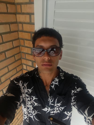 A man wearing sunglasses and a black button-down shirt with a white pattern looks down at the camera. He has short dark hair and is positioned outdoors, as indicated by the brick wall and ceiling visible in the background. The man has visible tattoos on his neck. The angle suggests the picture was taken from a low perspective, looking upward at him.