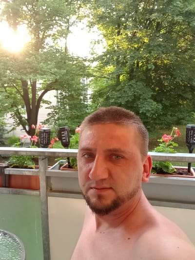 A close-up, waist-up selfie of a man is taken from his perspective as he looks directly at the camera. He has a short, buzzed haircut and a goatee. His eyes are blue-green, and his skin is fair. He is shirtless, and his shoulders and upper chest are visible.
The background suggests an outdoor setting, possibly a balcony or patio. In the mid-ground, a railing is visible, with potted plants containing pink flowers and green foliage. Two solar-powered garden lights are also present. Behind the railing, lush green trees with dappled sunlight filtering through their leaves dominate the scene. The sunlight suggests it is either early morning or late afternoon. The environment appears to be residential, with hints of buildings visible through the trees. The overall atmosphere is relaxed and natural. The provided location context of Chemnitz, Germany, places the scene within a European urban or suburban environment. No visible text is present in the image.