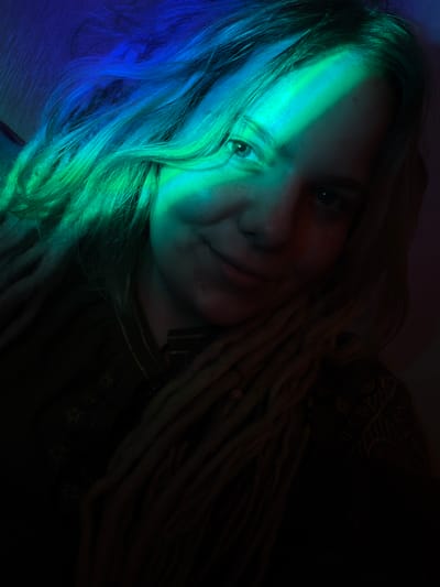 This is a medium shot, indoor, selfie photograph of a young woman with dreadlocks. She is looking at the camera and smiling slightly. Her face is illuminated by dramatic lighting, with a diagonal band of bright green and blue light across her forehead and cheek. The rest of her face and hair is in shadow, with some hints of warm reddish-brown light on the right side. She is wearing a dark-colored garment with some embroidered patterns visible on the left side of her chest. The background is dark and indistinct. There is no indication of the specific location within the city of Vostochny District, Russia.