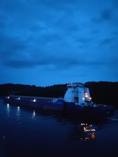 A barge is being pushed by a tugboat on a dark body of water at dusk. The tugboat is white with orange and red lights, and the barge is dark blue. Both vessels are moving towards the right of the image. The water reflects the lights of the tugboat. The background consists of a dark, tree-covered hillside against a deep blue sky with clouds. The location is identified as Yalosar', Russia.