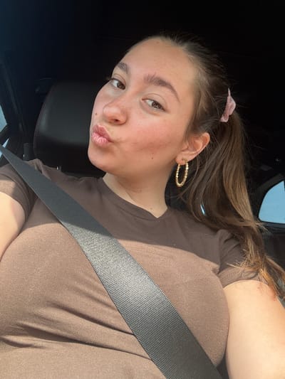 A young woman is taking a selfie in a car. She is wearing a brown t-shirt and a seatbelt. Her hair is brown and pulled back in a ponytail. She has hoop earrings and a pink scrunchie in her hair. She is puckering her lips. The background is dark, but appears to be inside a car. The image is taken in Polsko Padarevo, Bulgaria.