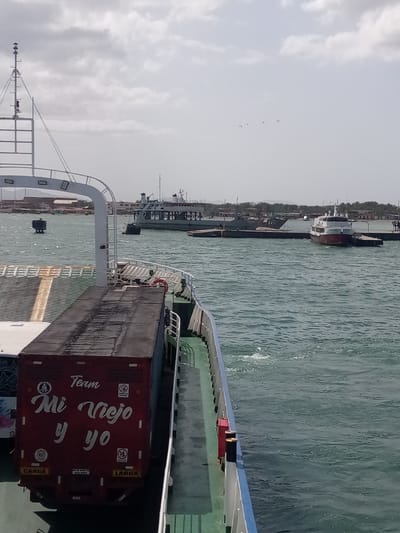 In Punta de Piedras, Venezuela, a ferry is docked or moving slowly in a body of water under a cloudy sky. On the deck of the ferry, a red trailer with white lettering is visible. The lettering reads "Team Mi Viejo y yo" and includes circular logos with "80" and other symbols. The trailer is positioned on the right side of the image, obscuring a large portion of the ferry's deck. In the background, other vessels, including a larger ferry or ship and smaller boats, are present in the water. Some landmass with buildings and vegetation is visible in the distance, suggesting a coastal or harbor area. The water appears choppy, indicating some movement or wind.