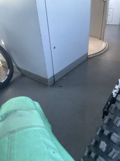 The image depicts an indoor scene with a grey speckled floor. In the foreground, a person wearing green pants is seated, partially visible. To the left, a bicycle wheel with a black tire and a white rim is positioned. In the background, there is a large, light-colored structure, possibly a wall or cabinet, with a metallic base. To the right of this structure, a circular entrance with a curved metal edge leads to another area. Further in the background, a white door with a small rectangular panel is visible on the right side. The lighting suggests an overhead light source. The setting appears to be a public indoor space, potentially a transit station or a building with distinct architectural features. The location in Merichleri, Bulgaria is not visually evident from the image itself.