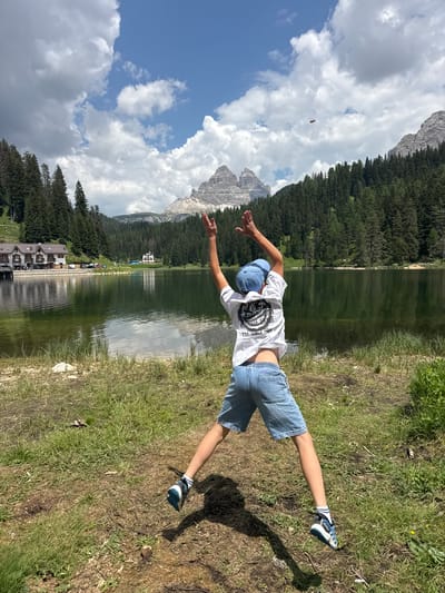A young person is captured mid-action, striking a dynamic pose with their arms outstretched as if flying, against a backdrop of a serene lake reflecting the sky and surrounding mountains. They are wearing a white t-shirt with a graphic print, denim shorts, a light blue baseball cap, and athletic shoes. The scene is set on the grassy bank of a lake, with a dense forest of evergreen trees lining the opposite shore, leading up to majestic, rocky mountains under a partly cloudy sky. The sun appears to be high in the sky, casting a distinct shadow on the grass, suggesting it is daytime, likely midday or early afternoon. The weather seems pleasant, with a mix of sunshine and clouds. In the distance across the lake, buildings are visible, hinting at a developed area or a resort. The overall mood is one of freedom and joy, amplified by the person's energetic pose and the beauty of the natural landscape. The text on the person's shirt is not clearly legible, but appears to be a bold graphic design with some stylized lettering. The location is identified as Auronzo di Cadore, Italy.