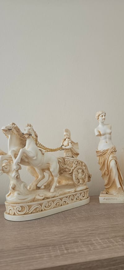 The image displays two decorative statues on a wooden shelf against a plain, light-colored wall. The statue on the left depicts a chariot pulled by two horses, with a figure in ancient Greek attire driving. The chariot has intricate detailing on its wheels and base, adorned with swirling patterns. The horses are white and appear to be in motion. The statue on the right is a partial depiction of a female figure, also in a classical style, draped in fabric. The base of this statue has Greek writing on it, which appears to read "Αφροδίτη" (Aphrodite). The overall impression is of a display of classical-themed decorative items. The image was taken in Hévíz, Hungary.