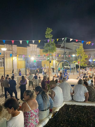 The image depicts an outdoor evening event in Olhão, Portugal, likely a festival or market, with a stage set up for a musical performance. Colorful triangular flags are strung across the scene, decorating the area between buildings. A band is visible on the stage, illuminated by spotlights, with a banner above them reading "LAGAR FREGUESIA MONCARAPACHO". A diverse crowd of people are gathered, some standing and observing the stage, while others are seated on benches or at tables. A unique blue circular apparatus, possibly part of an amusement or exhibit, is present in the mid-ground. In the background, several buildings with light-colored facades and balconies line the square, and trees with lush green foliage are scattered throughout the scene. The overall atmosphere appears lively and communal, with attendees engaged in socializing and enjoying the entertainment.