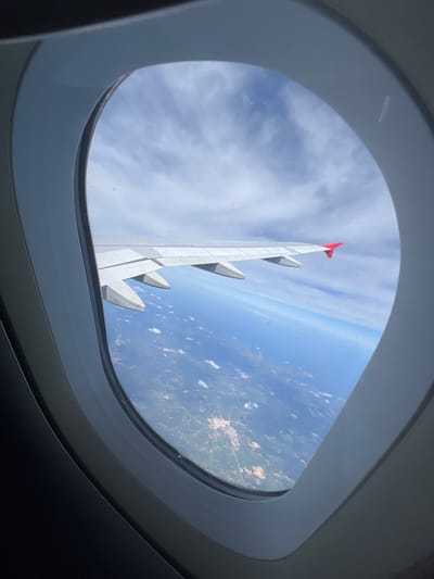 The image presents an aerial view captured from inside an aircraft through a window, whose grey frame partially encloses the scene. The grey right wing of the airplane, featuring multiple flap fairings on its underside and a prominent red wingtip, spans diagonally across the middle ground. Above the wing, the sky exhibits scattered white stratocumulus clouds against a clear blue expanse. Below the wing, the landscape transitions from a patchwork of green vegetation and lighter, developed areas containing small structures, consistent with the city of Maó, Spain, on the island of Menorca. A distinct, deep blue body of water, representing the Mediterranean Sea, occupies the background, meeting the land at the horizon.
