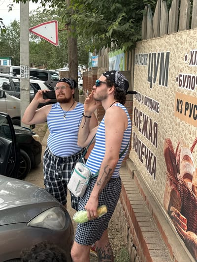 Two men are standing next to a brick wall with a large advertisement for baked goods. Both men are wearing blue and white striped tank tops, black and white checkered shorts, and black bandanas. The man on the left is wearing glasses and holding a black smartphone to his ear. He has a beard and a pendant with a tooth-like charm around his neck. The man on the right has sunglasses on his head and is smoking a cigarette. He is carrying a light-colored messenger bag and holding a loaf of bread wrapped in plastic. The advertisement behind them features images of bread and text in Russian. In the background, there are cars, trees, and a red and white triangular road sign. The ground is a mixture of dirt and pavement. The overall scene suggests an outdoor urban or suburban environment. The location is identified as Zhemchuzhny, Russia.