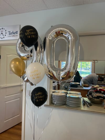 The image shows a cluster of balloons, including two large silver foil balloons shaped like the number "80", along with three smaller latex balloons. Two of the latex balloons are black with gold lettering that reads "Happy Birthday to You", and one is white with the same gold lettering. There is also a gold latex balloon. The balloons are tied with silver ribbons.
In the background, there's a glimpse of a person standing near a countertop with stacked plates and utensils. The reflection in a mirror shows a window with an outdoor view of trees. The location is likely indoors, as evidenced by the presence of a door and what appears to be a wall or partition. There is a Tarkett clock that indicates it may be in the city of Kelowna, Canada.