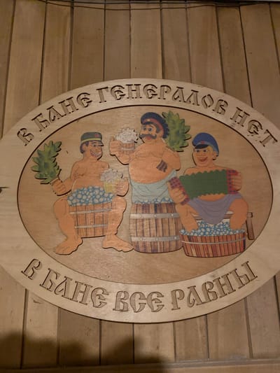 This is a circular wooden plaque with carved and painted illustrations and text. The central image depicts three men in a bathhouse setting. The top man, wearing a blue cap and a purple loincloth, is playing an accordion. He is positioned above two wooden barrels. Below him, another man, with a dark beard and blue cap, is holding a venik (a bath broom made of branches) and a mug. He is also partially enclosed by a wooden barrel. The bottom man, also with a dark beard and blue cap, is reaching for a venik and holding a mug. He is also surrounded by a barrel. The men are depicted with smiles, suggesting a cheerful atmosphere. The setting appears to be a traditional Russian banya, a type of steam bath, as indicated by the veniks and the presence of barrels, which are often used for water and heating. The wood grain of the plaque is visible, and it is mounted on a wooden wall composed of horizontal planks.
The text inscribed in Russian along the outer edge of the circle reads: "В БАНЮ ГЕНЕРАЛОВ НЕТ" on the left side, which translates to "No generals in the banya." On the right side, the text reads: "Я В БАНЕ ВСЕ РАВНЫ" which translates to "We are all equal in the banya." This proverb emphasizes the egalitarian nature of a banya. The carving and painting are stylized, with bold outlines and vibrant colors. The lighting in the image is warm, suggesting an indoor environment. The location context provided is Ranchpar, Armenia.
