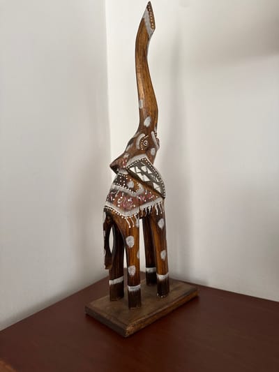 The image displays a handcrafted wooden elephant sculpture positioned indoors. The specific city location within AMCO, Area Metropolitana Centro Occidente, Colombia, is not discernible from the visual information provided. The sculpture, crafted from dark brown wood, features an elongated, stylized elephant standing with its trunk raised and curved upwards. It is adorned with white painted patterns, including dots on its legs and trunk, stripes on its lower legs, and a geometric motif on its back/saddle area. Additional swirling patterns in white and reddish-maroon are present on the body. The elephant stands on a flat, rectangular base of lighter brown wood, exhibiting minor wear. This base rests on a polished, dark reddish-brown wooden surface, likely a table or shelf, which occupies the lower portion of the frame. The background consists of a plain white wall, forming an interior corner on the left side of the image. No people, actions, or interactions are visible.