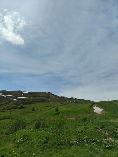 The image depicts a scenic view of a grassy mountainside with patches of snow. The sky is partly cloudy, with white clouds scattered against a pale blue background. In the mid-ground, a ski lift with poles and cables is visible, indicating a ski resort. The terrain is uneven and slopes upwards towards the peak, which is partially covered in snow. The foreground is dominated by lush green grass, with some bushes and smaller plants. The scene suggests a natural landscape in a mountainous region, possibly during the spring or early summer when snow still lingers on higher elevations. The location is identified as Bakuriani, Georgia, a well-known mountain resort. There are no people or animals visible in the image. The overall atmosphere is serene and picturesque.