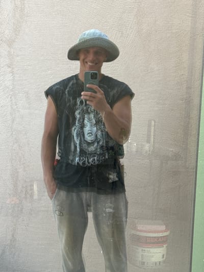 A man in his late 20s or early 30s, wearing a blue patterned bucket hat, a black t-shirt with a graphic print, and light grey sweatpants, is taking a selfie in a mirror. The man is smiling and holding a smartphone in his right hand. The t-shirt is sleeveless, exposing his muscular arms. The sweatpants have a logo on the left thigh. The background appears to be a textured wall, possibly exterior stucco, and a green-tinted vertical element on the right. In the lower right corner, a white bucket with red and black labeling, including the word "BEKAME", is visible. The scene appears to be outdoors, possibly in a construction or renovation area, given the presence of the bucket and the man's attire. The image suggests an informal, casual setting. It is not possible to determine the specific location within the city of Pelinovo, Montenegro.