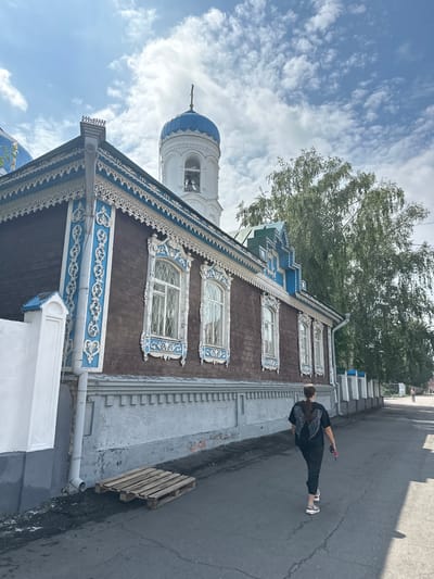 A woman with dark hair, wearing a black t-shirt with white wings on the back and patterned leggings, walks away from the camera down a paved street. She is on the right side of the street, casting a shadow onto the pavement. To her left is the exterior of a traditional wooden Russian building, painted dark brown with ornate white and blue trim around the windows and roofline. The building has multiple windows with decorative frames. A blue-domed bell tower rises behind the building, topped with a cross. Adjacent to the building's foundation is a white, stuccoed wall with a blue cap. A metal drainpipe runs down the side of the building. On the ground in front of the building is a wooden pallet. The street is lined with a white fence with blue accents. Behind the building and fence, a large, leafy green tree is visible. The sky is blue with scattered white clouds. This scene is located in the city of Nogornyi, Russia.