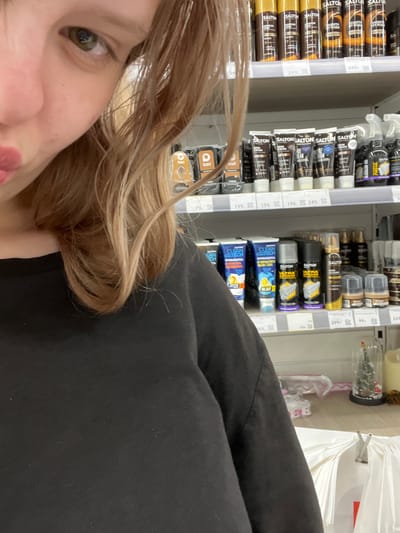 A close-up shot captures the left side of a young woman's face, focusing on her eye and lips, as she looks towards the camera. Her light brown hair is parted on the left and falls across her shoulder. She is wearing a black long-sleeved shirt. The background consists of store shelves displaying various shoe care products, including bottles, tubes, and sprays. Some of the product labels are visible, such as "SALTON" and "CLEAN TECH." Price tags are also present on the shelves. The overall environment appears to be a retail setting, likely a shoe care or cosmetics section of a store. The image's context suggests it was taken in a store located in Kokkorevo, Russia.