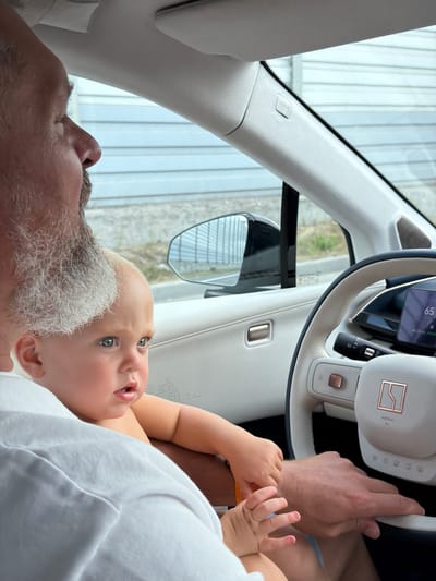 An adult, likely a male, with graying facial hair is in the driver's seat of a modern car. He is wearing a white shirt. A young child, a baby, is seated on his lap, facing towards the right side of the frame. The baby has fair skin and blue eyes. The interior of the car is visible, including a steering wheel with controls and a digital display showing "65". The background outside the car's window appears to be a light-colored building with horizontal siding, suggesting an urban or suburban environment. The image is taken from a slightly elevated perspective, looking down and to the right.
